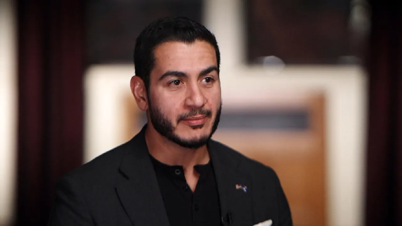 A bearded man in a dark blazer looks off-camera during an indoor interview.