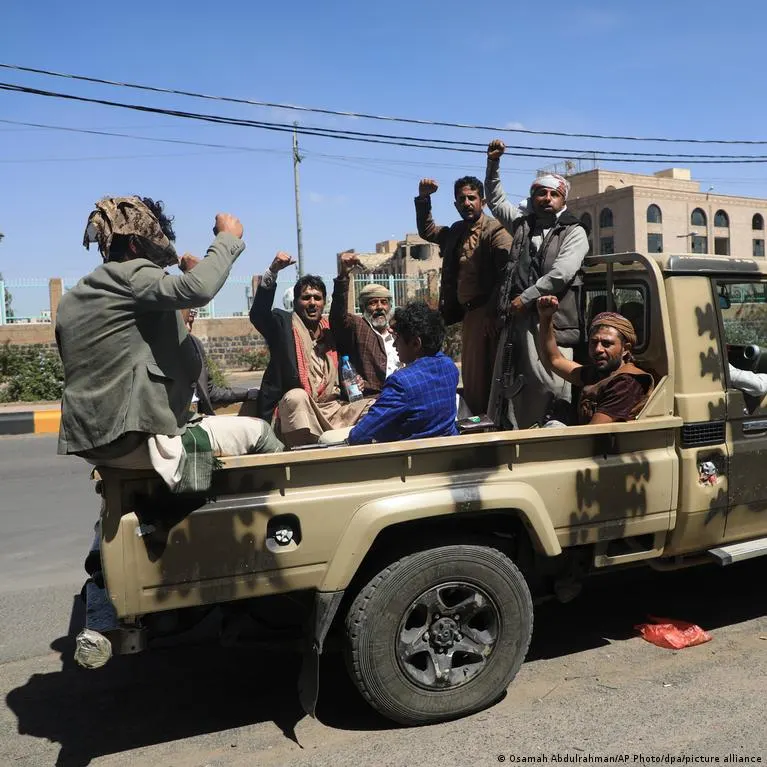 Armed men in a camouflage pickup truck raise fists and cheer on a city street.