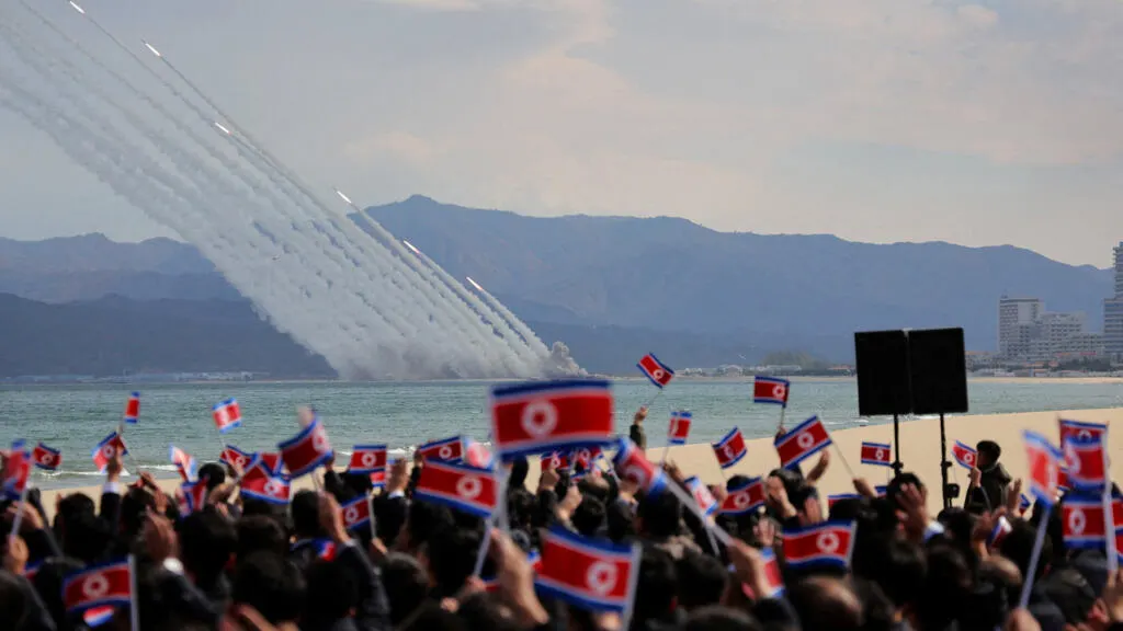 Crowd waving North Korean flags as smoke trails of rockets streak over a coastal mountain scene
