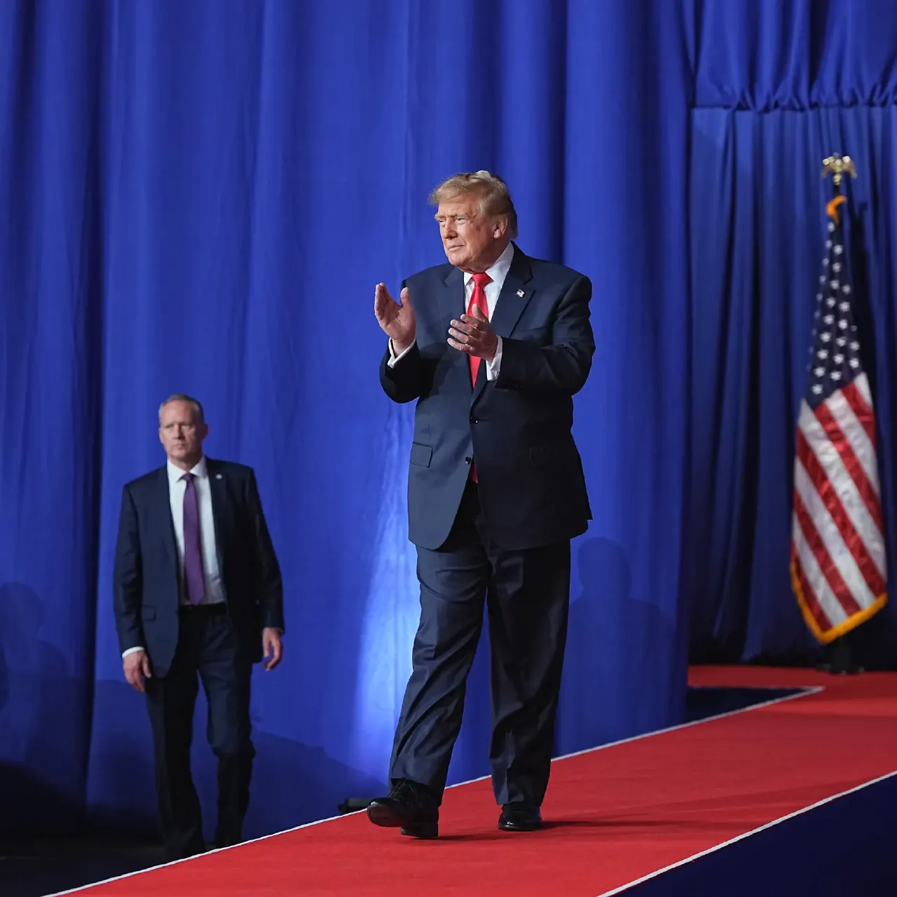 A man in a navy suit clapping on a stage with blue drapes and an American flag.