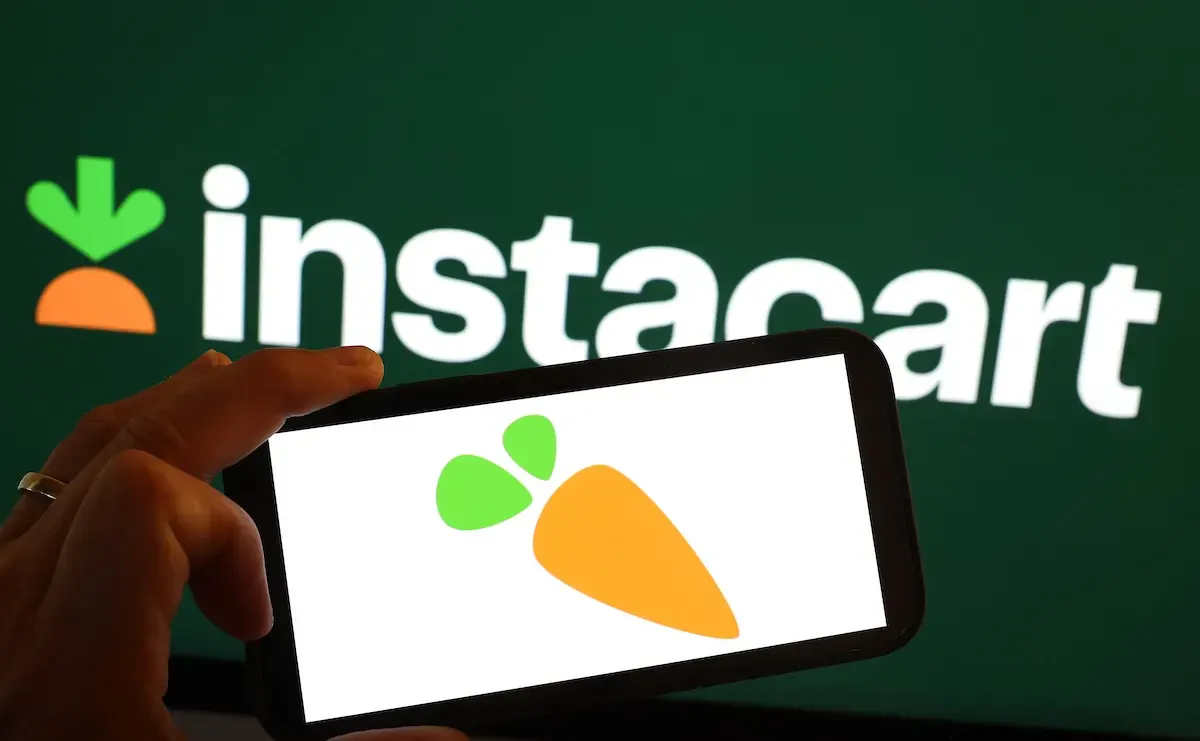 Hand holding phone showing Instacart carrot logo before large green Instacart wordmark backdrop.