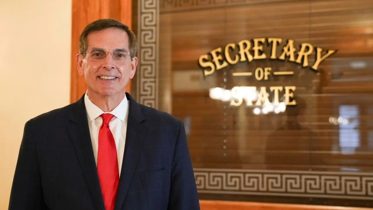 Smiling man in navy suit and red tie beside gold 'Secretary of State' office sign.