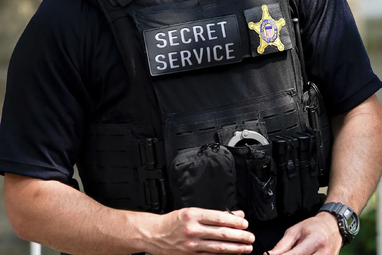 Close-up of a Secret Service officer's tactical vest with name patch, badge, and handcuffs.