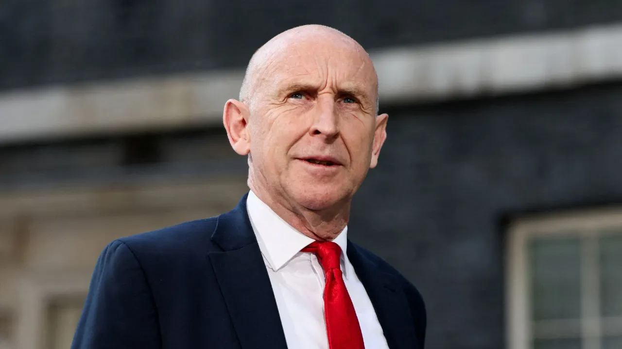 Bald man in dark suit, white shirt and red tie before a dark brick building.