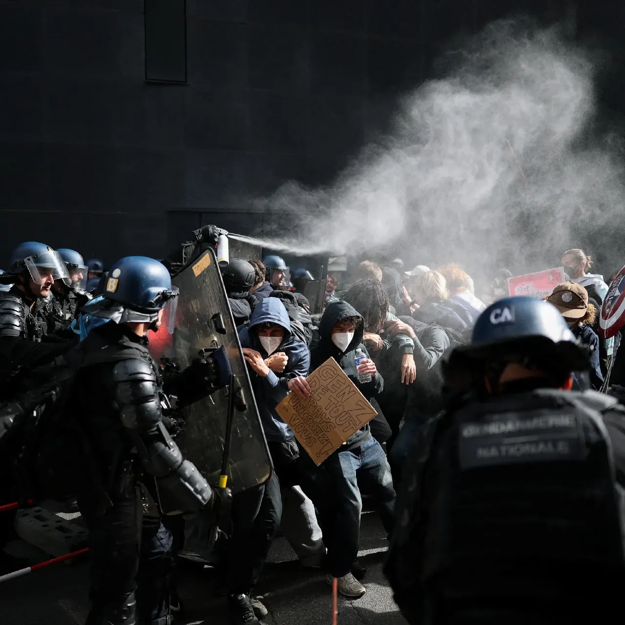 Riot police with shields face protesters as tear gas or spray drifts through the street