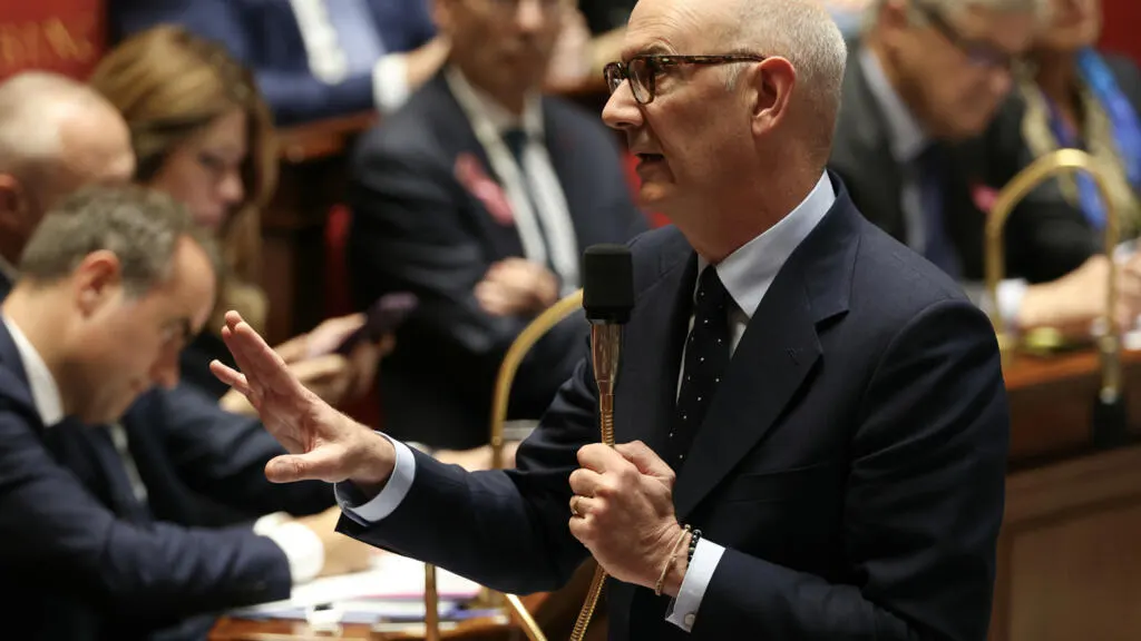 Man in dark suit speaking into microphone and gesturing in a legislative chamber.