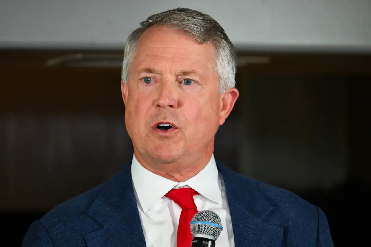 Older man with gray hair in navy suit, red tie, speaking into a microphone.