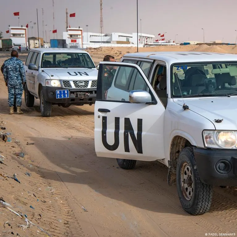 White UN vehicles on a sandy road at a desert base, with a uniformed guard and red flags.