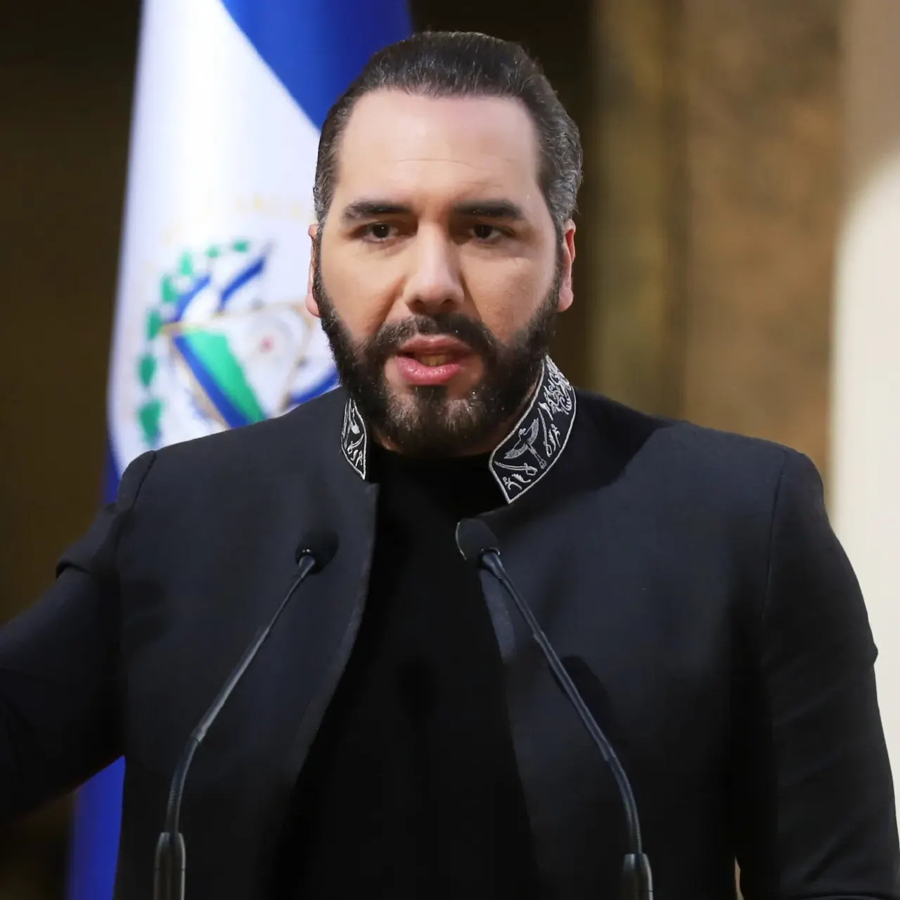 A bearded man in a black high-collared jacket speaks at a microphone before a national flag.