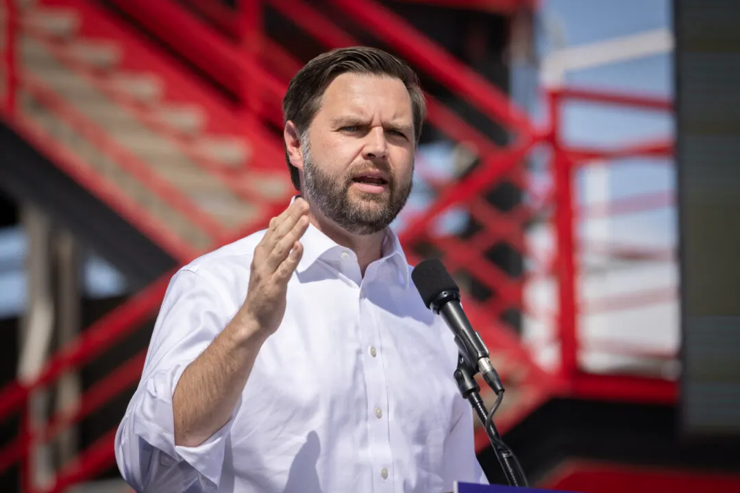 Man in white shirt gestures while speaking at a microphone before red metal stairs.