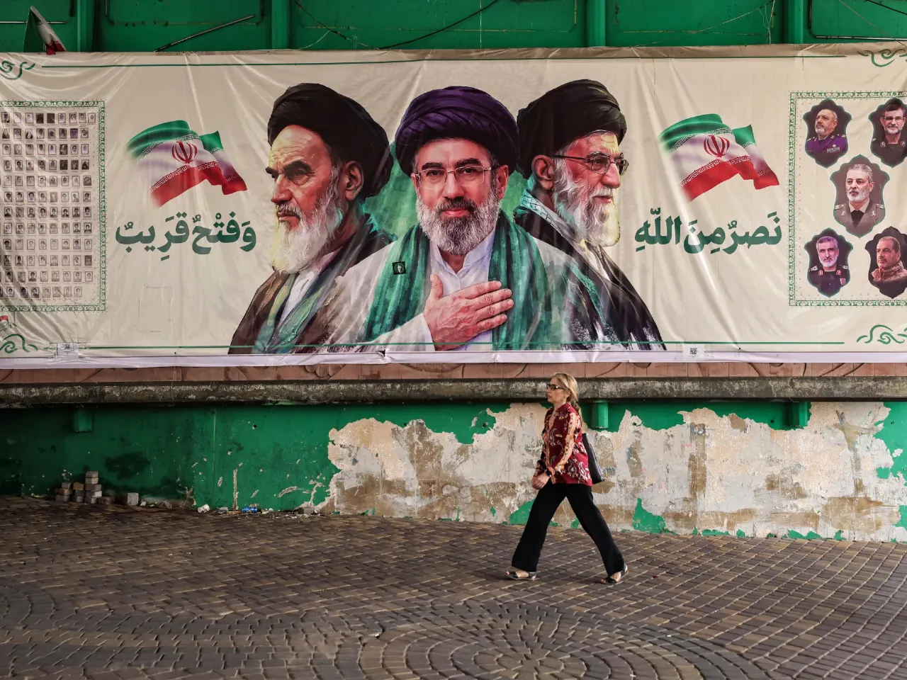 Woman walks past large street mural of three bearded clerics, Iranian flags, Arabic script, green wall.