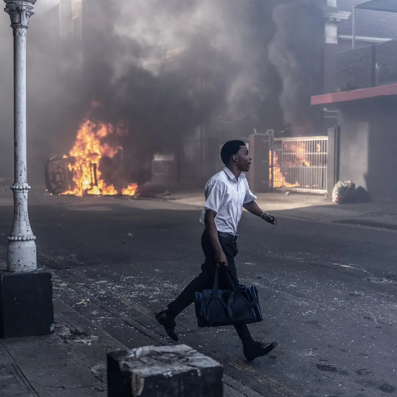 A young man in a white shirt walks past a burning fire on a smoky street.