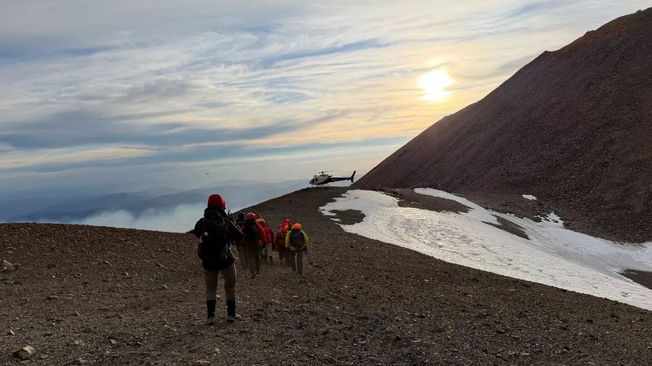 Rescuers hiking up a rocky mountain slope toward a helicopter at sunset near snow patches.