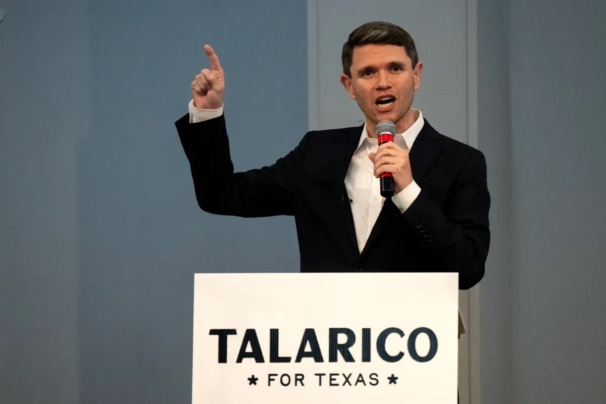 Man in dark suit speaking at podium with microphone, pointing up, "Talarico for Texas" sign