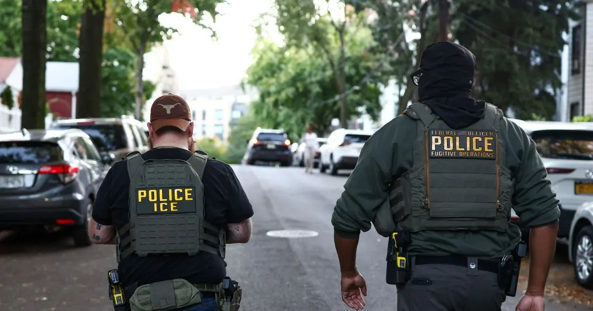 Two officers in tactical vests marked ICE and Fugitive Operations walk down a residential street.