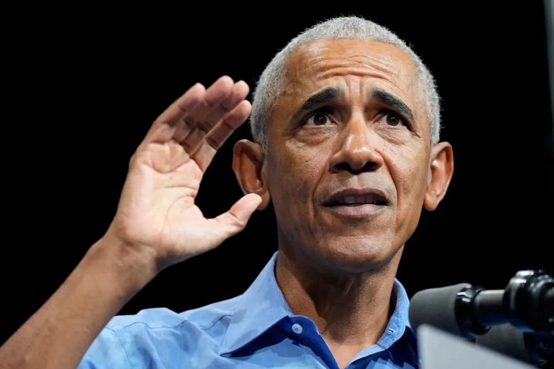 Older man in blue shirt gesturing with raised hand beside a microphone on a dark stage.