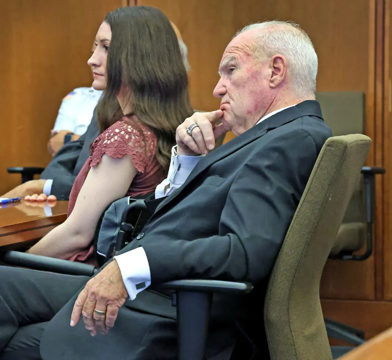 Older man in dark suit with hand at chin beside woman in courtroom seating.