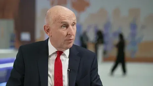 Bald man in dark suit and red tie speaking in a studio interview setting.