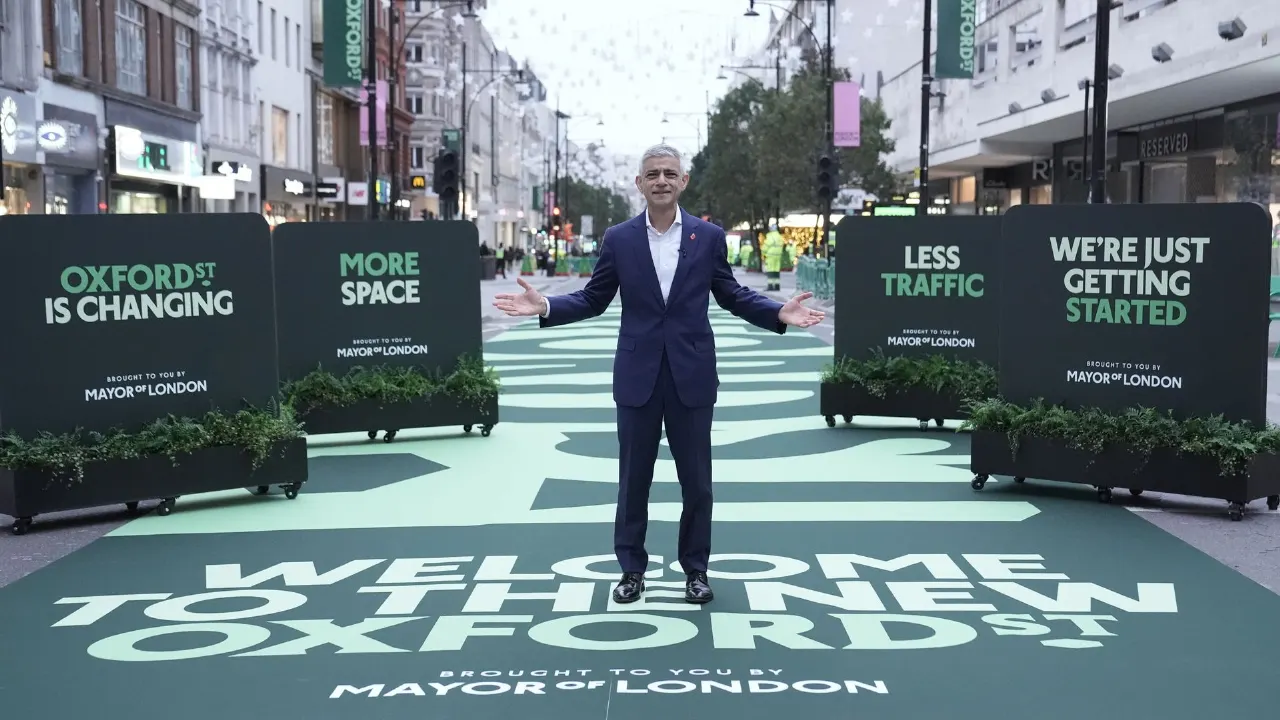 Man in suit arms outstretched on Oxford Street, with Mayor of London project signage and pedestrian street graphics.