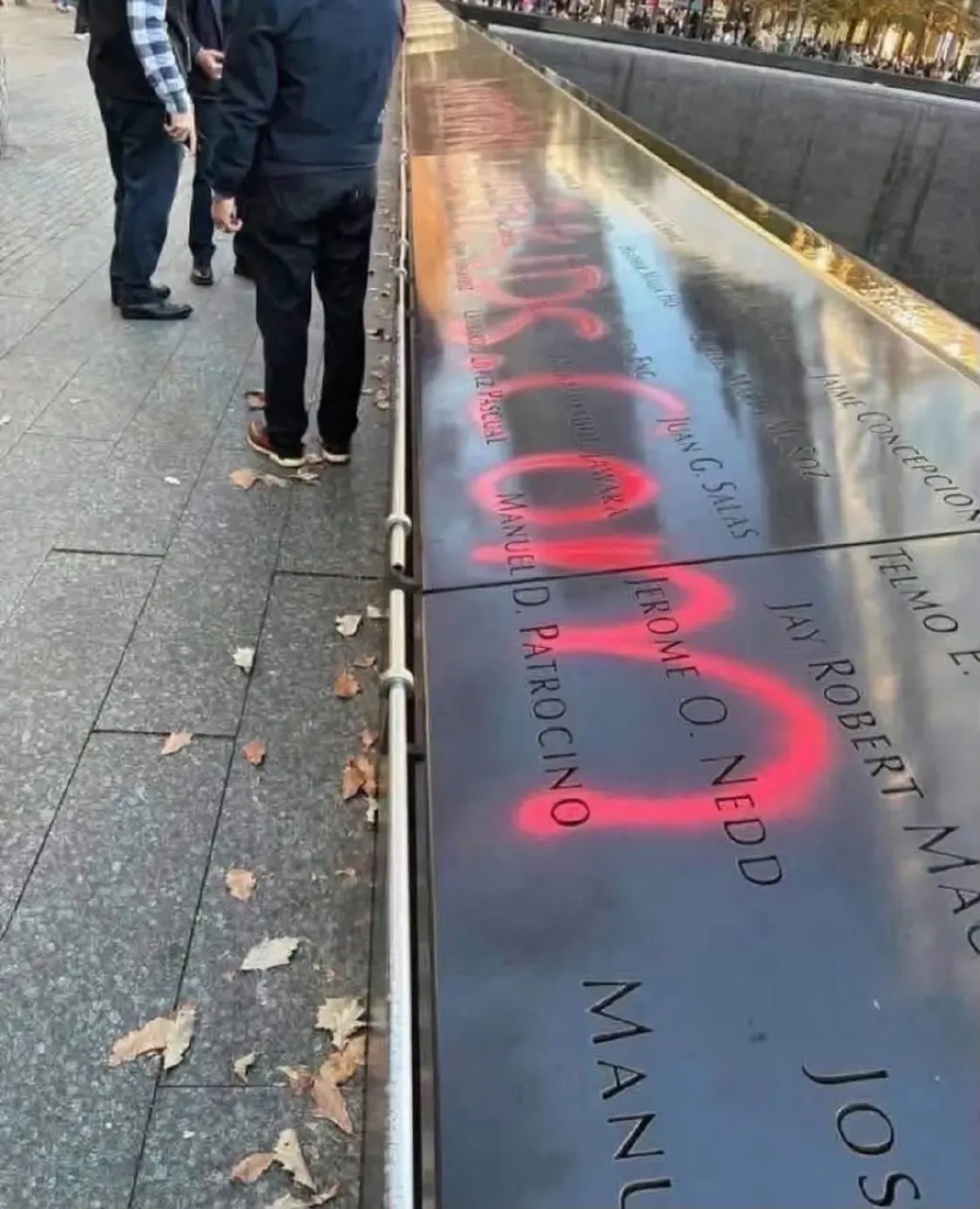 Red spray paint defacing names on the 9/11 Memorial reflecting pool edge, people standing nearby.