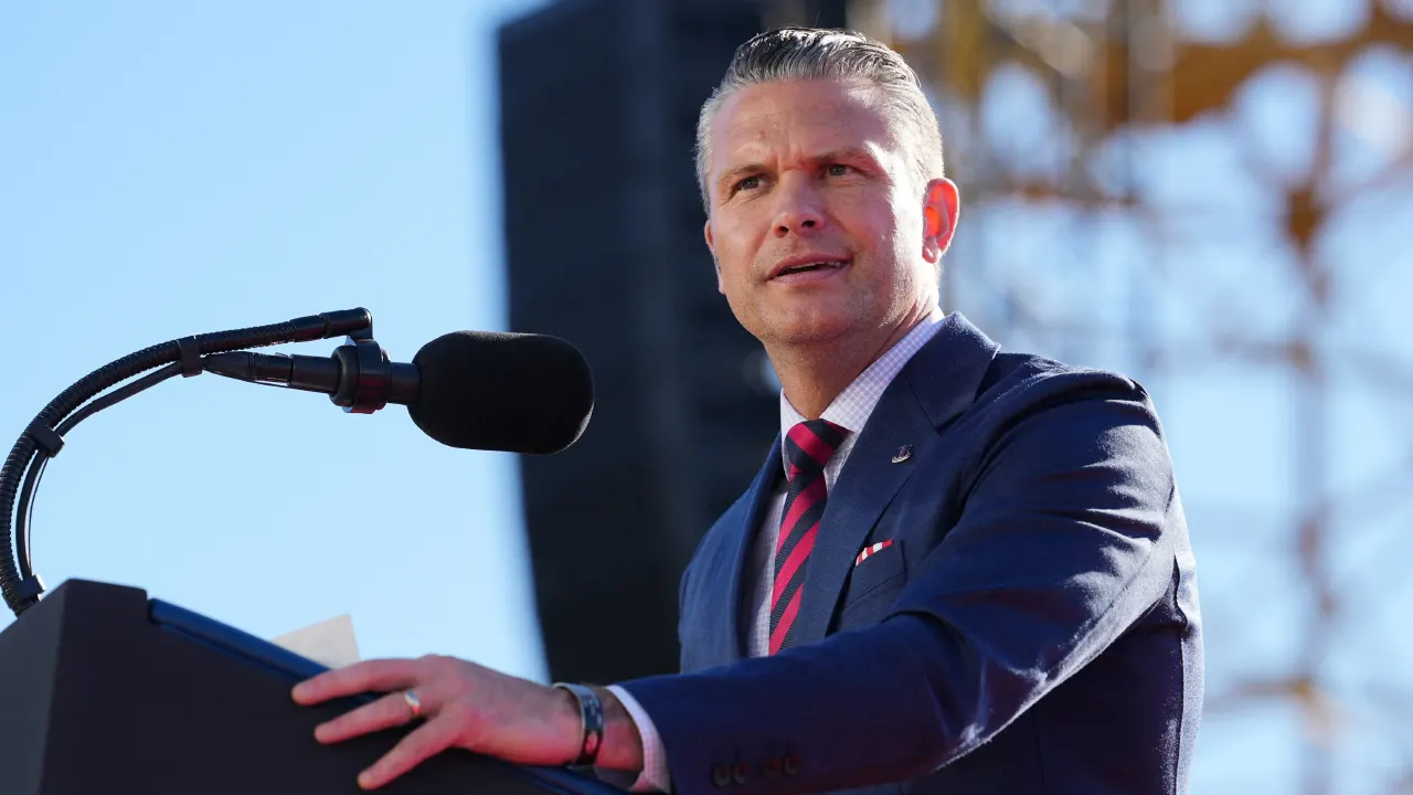 A man in a navy suit speaks at an outdoor podium with a microphone.