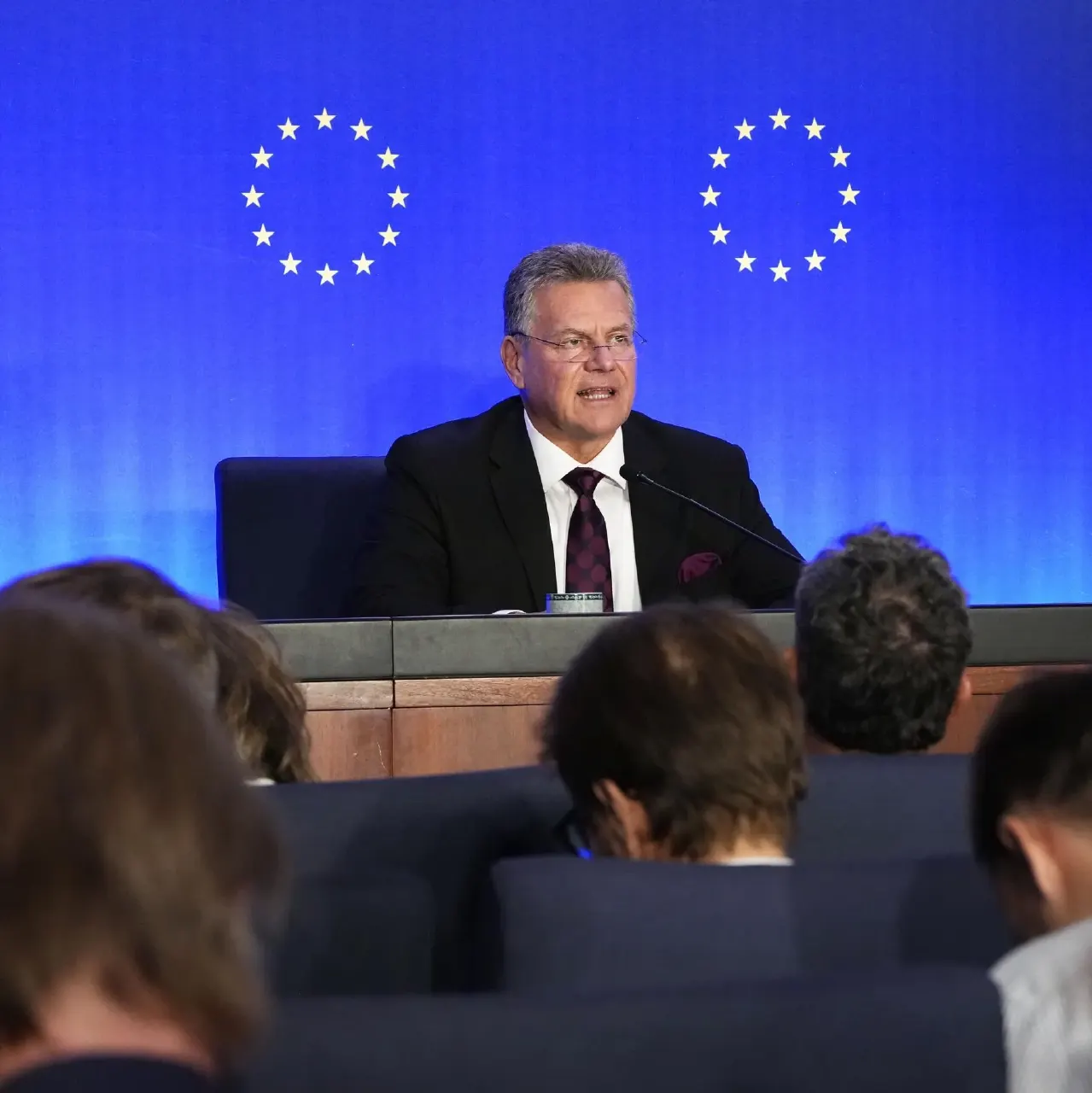 Man in dark suit speaks at press briefing before blue backdrop with EU star circles, audience in foreground.