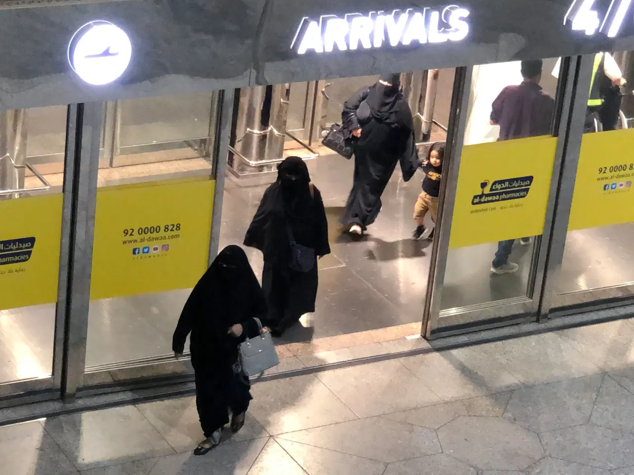 Women in black abayas walk through airport Arrivals doors, a pharmacy sign visible, elevated view.