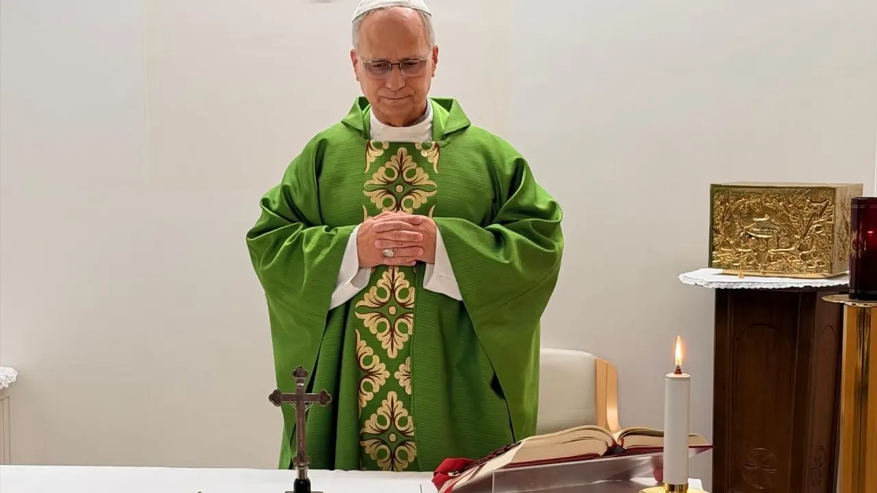 Clergyman in green vestments with hands clasped praying at altar with cross, candle, and book.