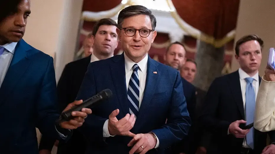 Man in dark suit and glasses speaking amid aides and reporters in a hallway.