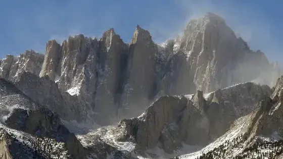 Snow-dusted jagged granite peaks under blue sky with wind-blown snow plumes.