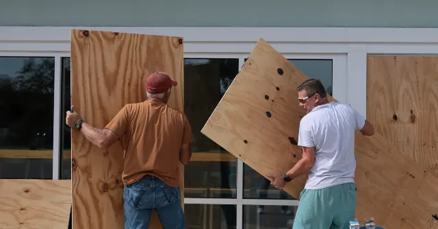Two men installing plywood storm shutters over the windows of a building.
