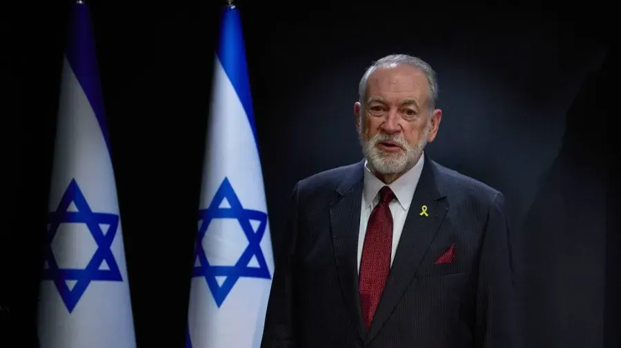 Man in dark suit and red tie stands before two Israeli flags against dark background.