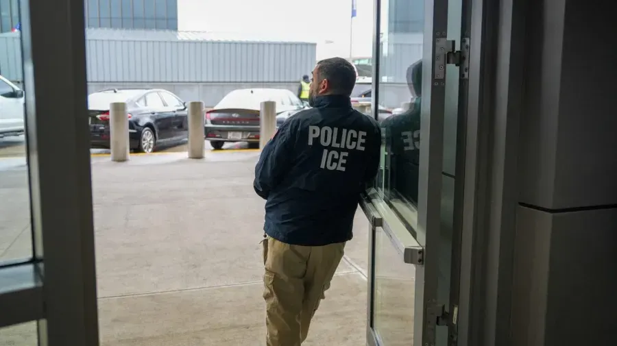 Person in jacket marked POLICE ICE walking through glass doorway, back to camera.