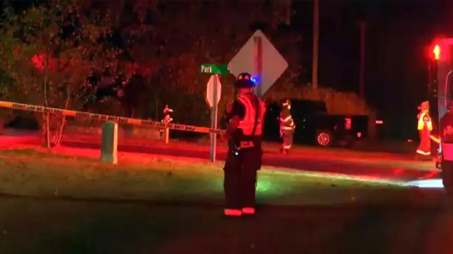 Firefighters at night behind police tape on a road, emergency lights glowing red.