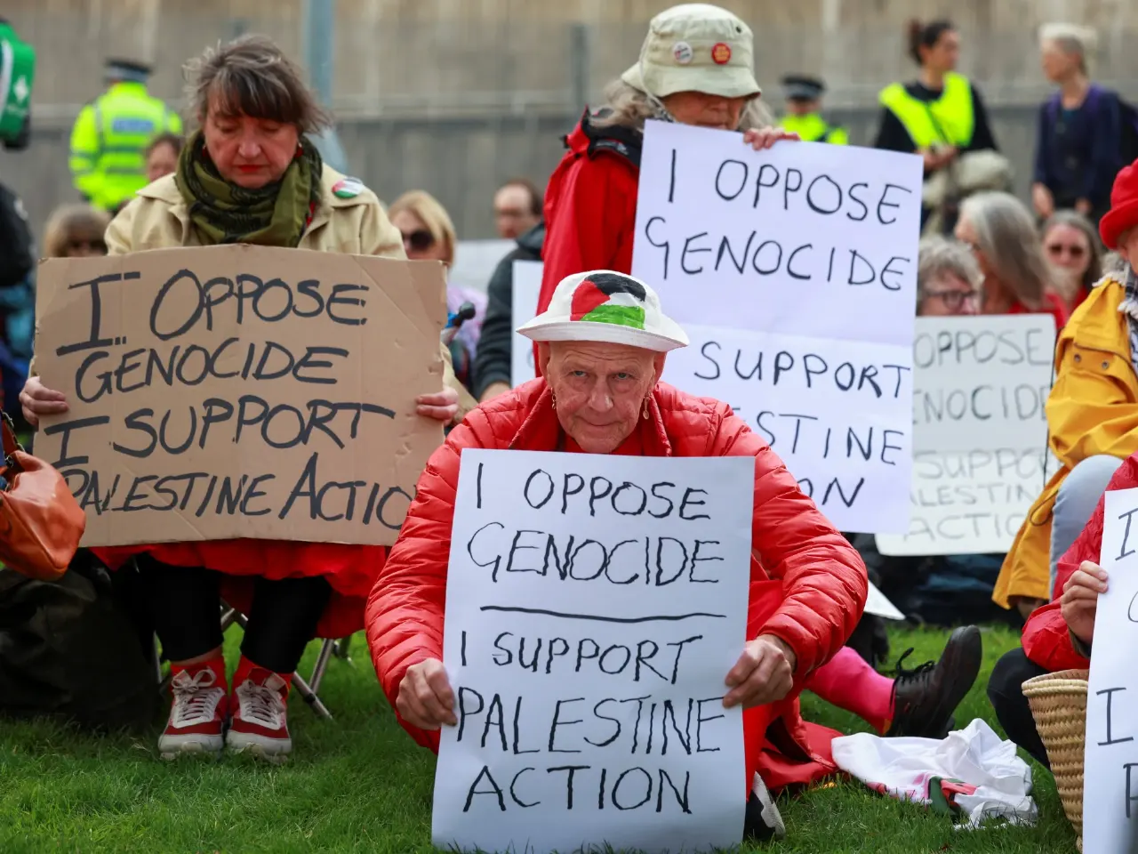 Protesters seated on grass holding handmade signs reading 'I oppose genocide, I support Palestine Action'.