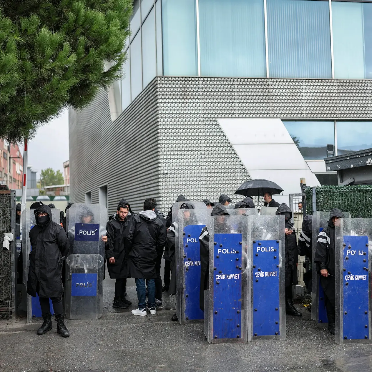 Black-clad police in hoods holding blue 'POLIS ÇEVİK KUVVET' riot shields outside a glass building.