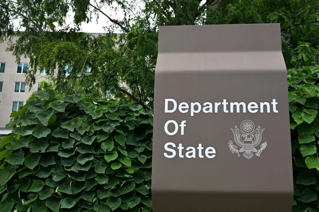 Brown sign reading Department of State with the Great Seal, in front of green foliage and a building.