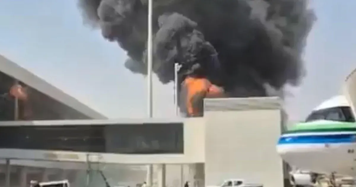 Thick black smoke and fire rising behind an airport terminal beside a parked aircraft.