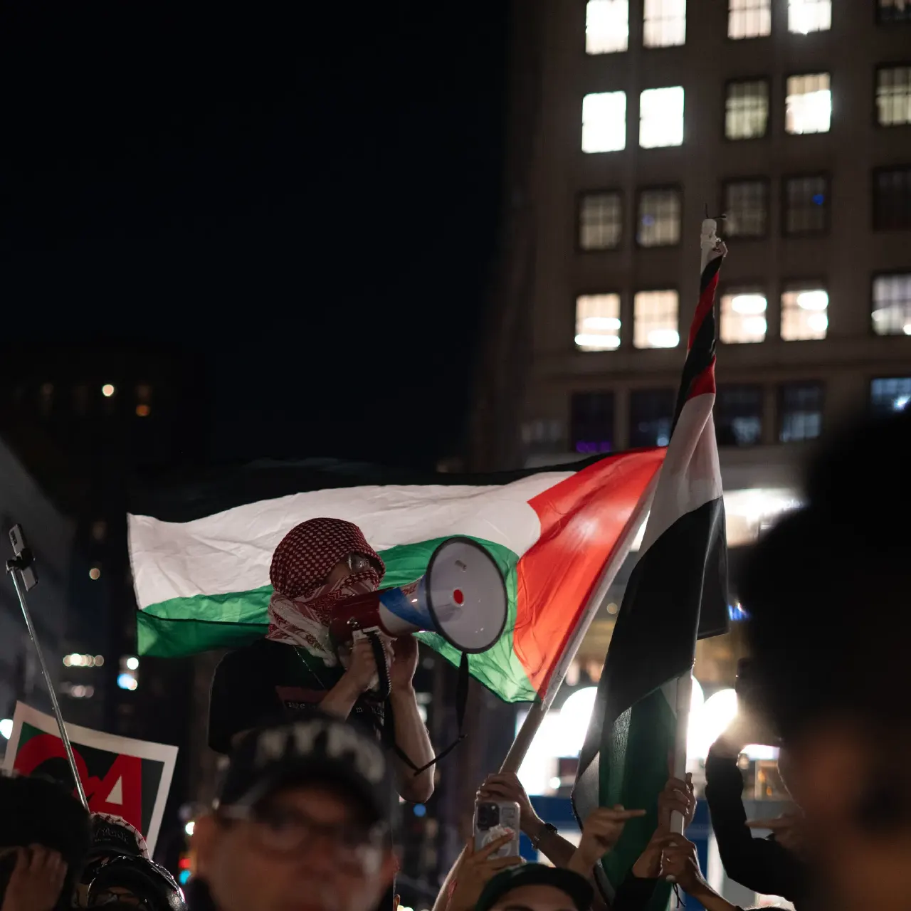 Night protest crowd with a person using a megaphone beneath large Palestinian flags, city buildings behind.