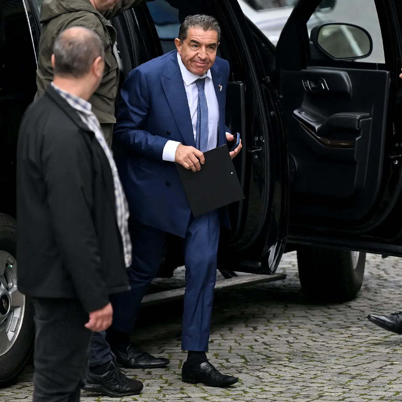 Man in navy suit exits black SUV on cobblestones, with two men beside him.