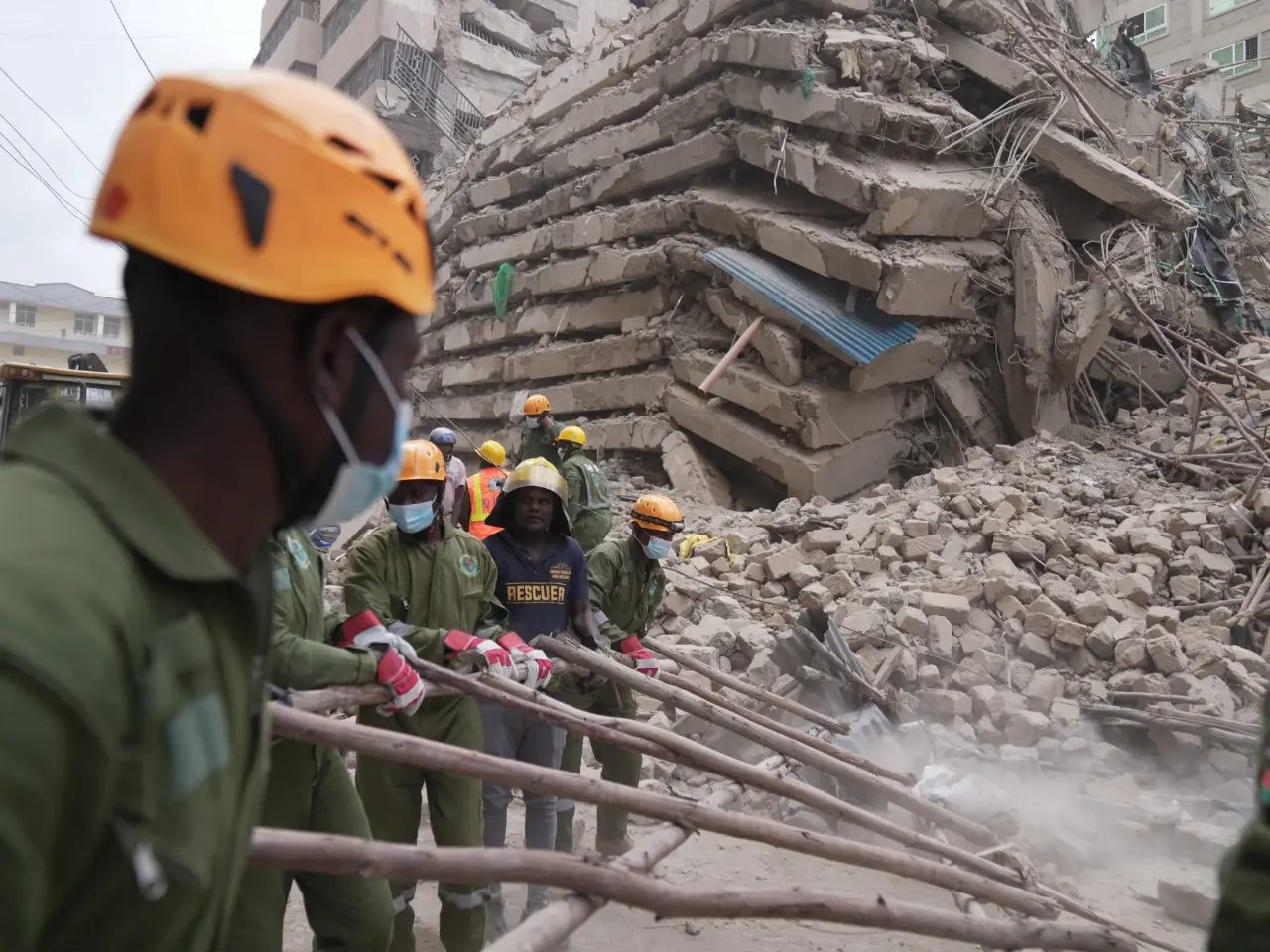Rescue workers in helmets and masks clear rubble from a collapsed multi-storey building.