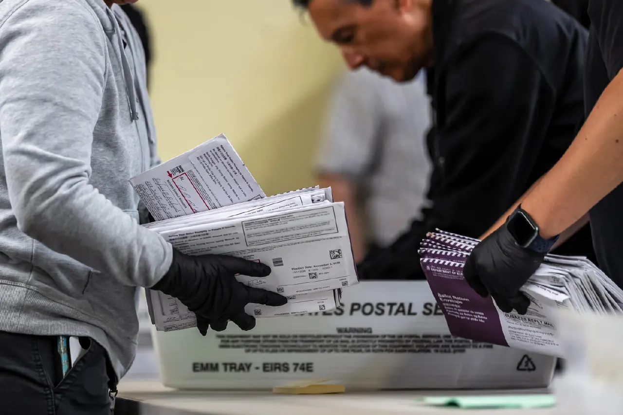 Gloved workers handle stacks of mail-in ballot envelopes and return packets over a Postal Service tray.