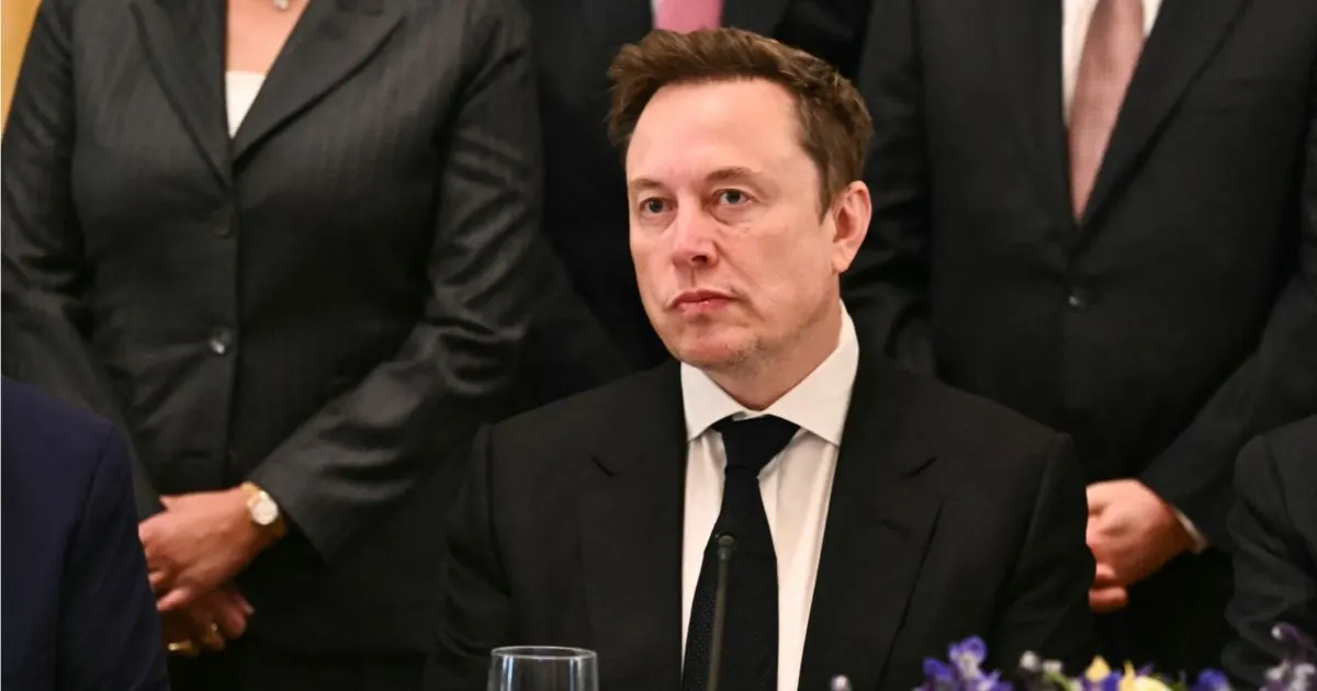 Man in dark suit and tie seated at a formal event, with standing people behind him.