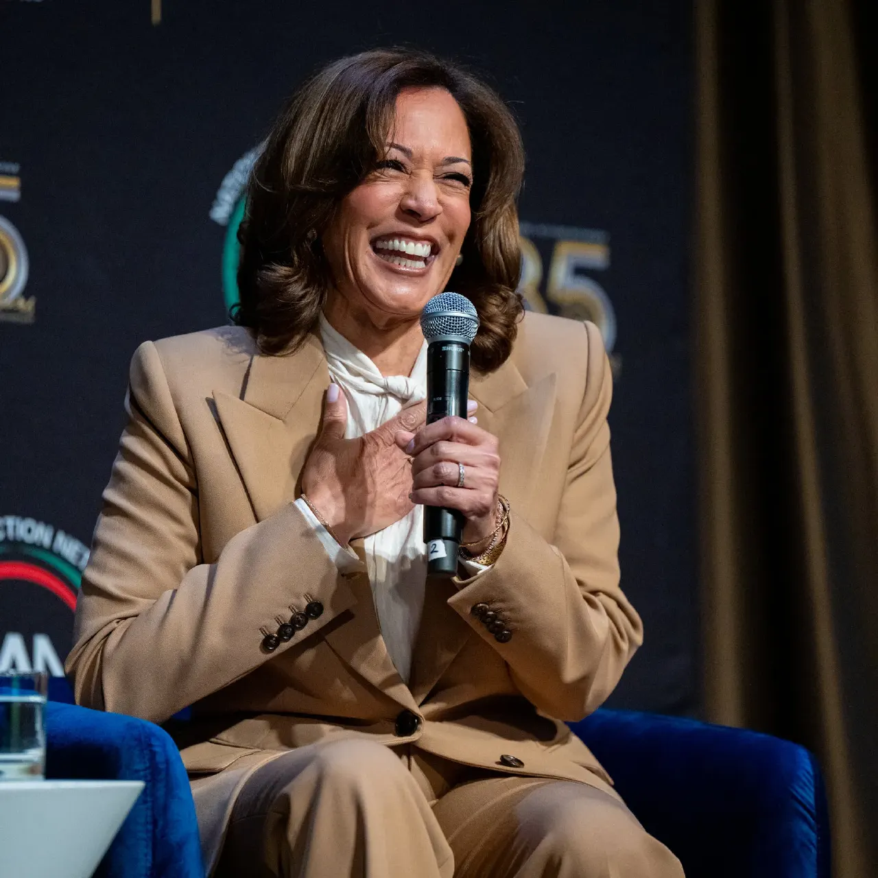 Smiling woman in tan suit holding a microphone while seated on a blue chair at an event.