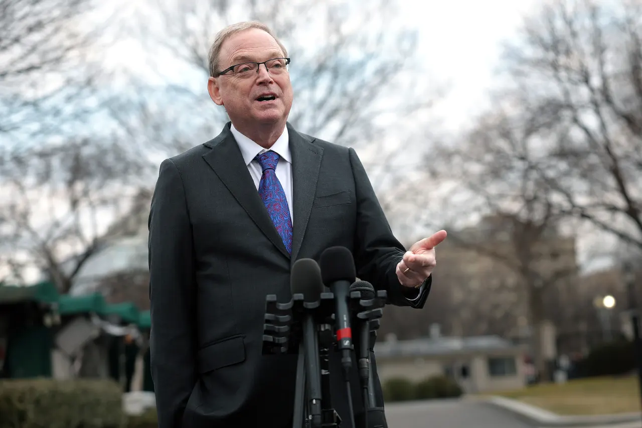A man in glasses and dark suit speaks at a cluster of microphones outdoors.