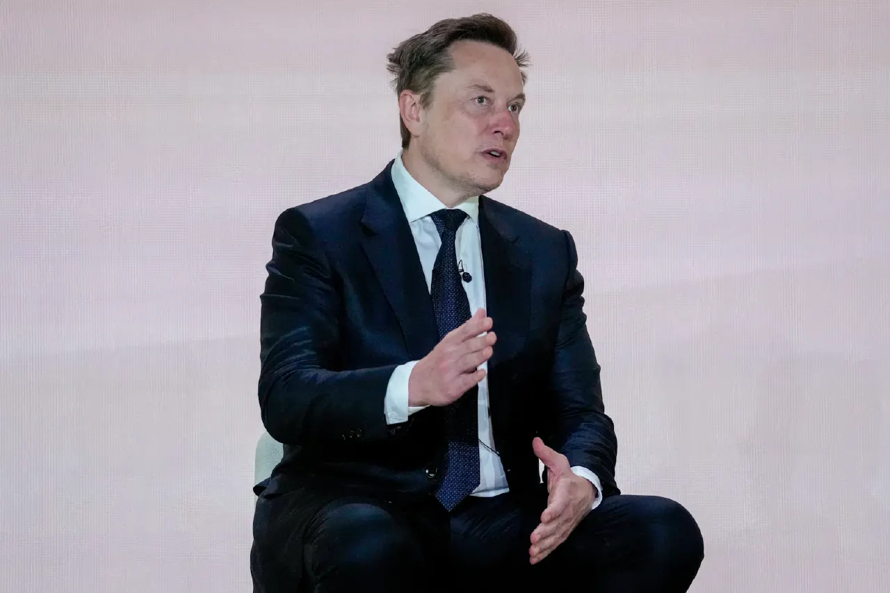 A man in dark suit and tie gestures while speaking, seated against a pale pink backdrop.