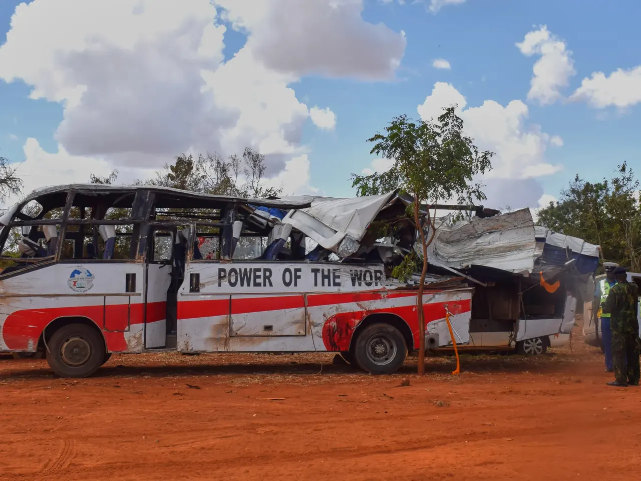 Crushed, roofless bus with 'Power of the Word' lettering on dirt road, officers standing nearby.