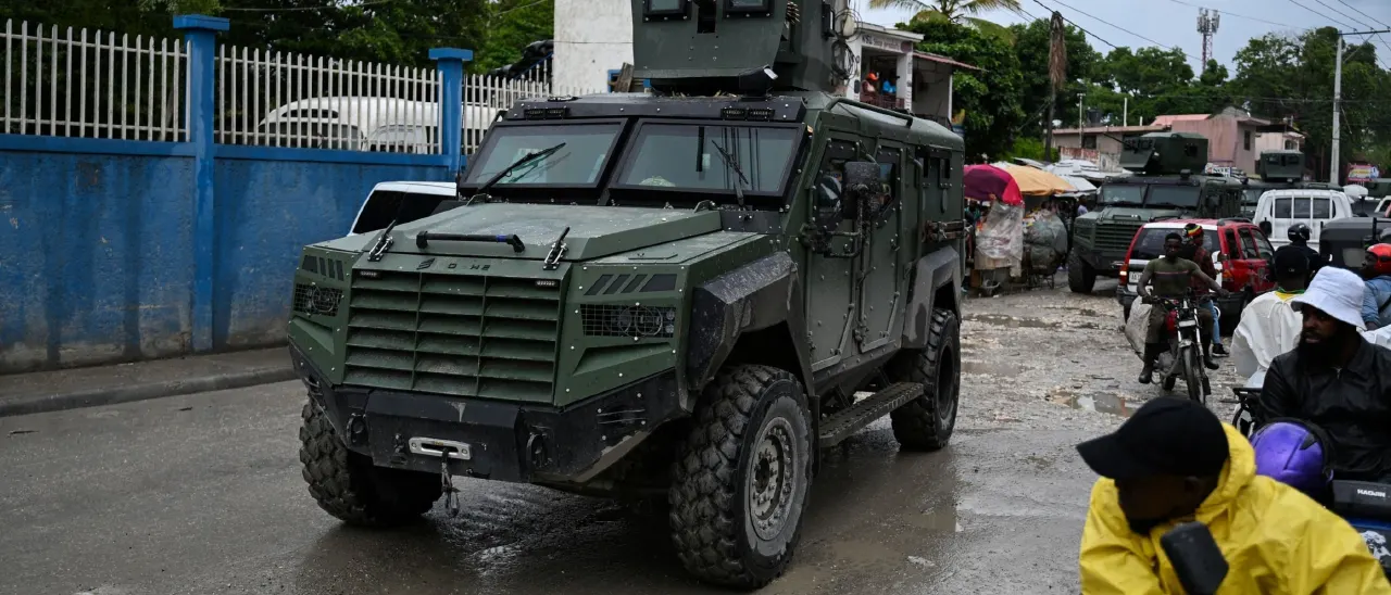 Armored military-style vehicle with a turret drives down a rainy street among pedestrians and motorcycles.