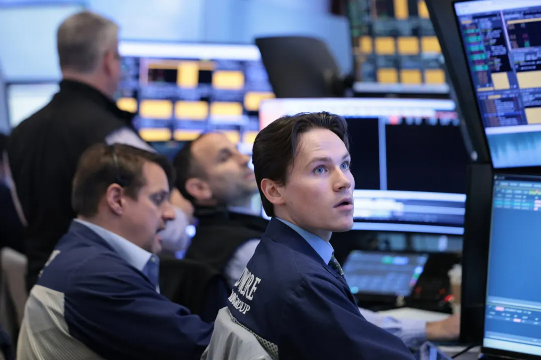 Traders in navy jackets watch multiple trading monitors on a busy financial trading floor.