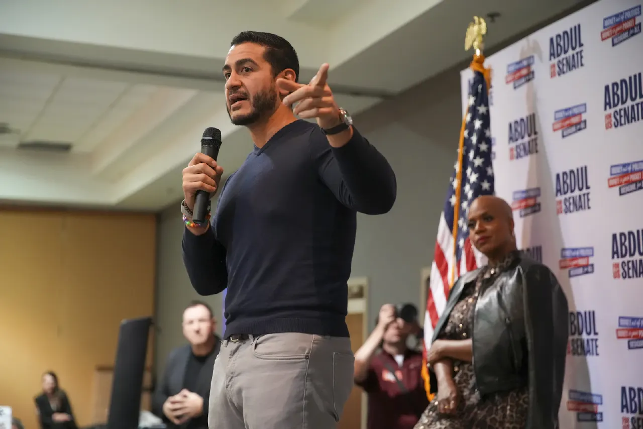 Man in navy sweater speaking into microphone at town hall, gesturing; campaign backdrop and US flag behind.
